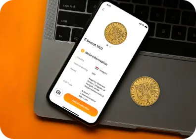 A smartphone on a laptop with a coin information card open on the screen; a gold coin lies next to it.